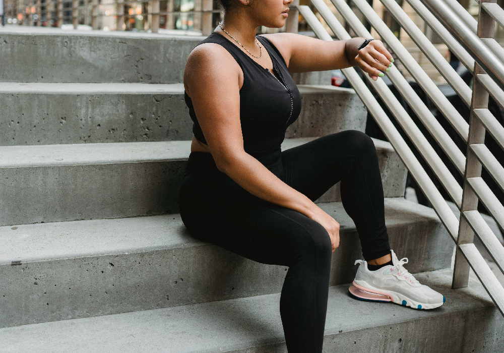 Woman in black activewear sitting on outdoor steps and checking her smartwatch after exercise.