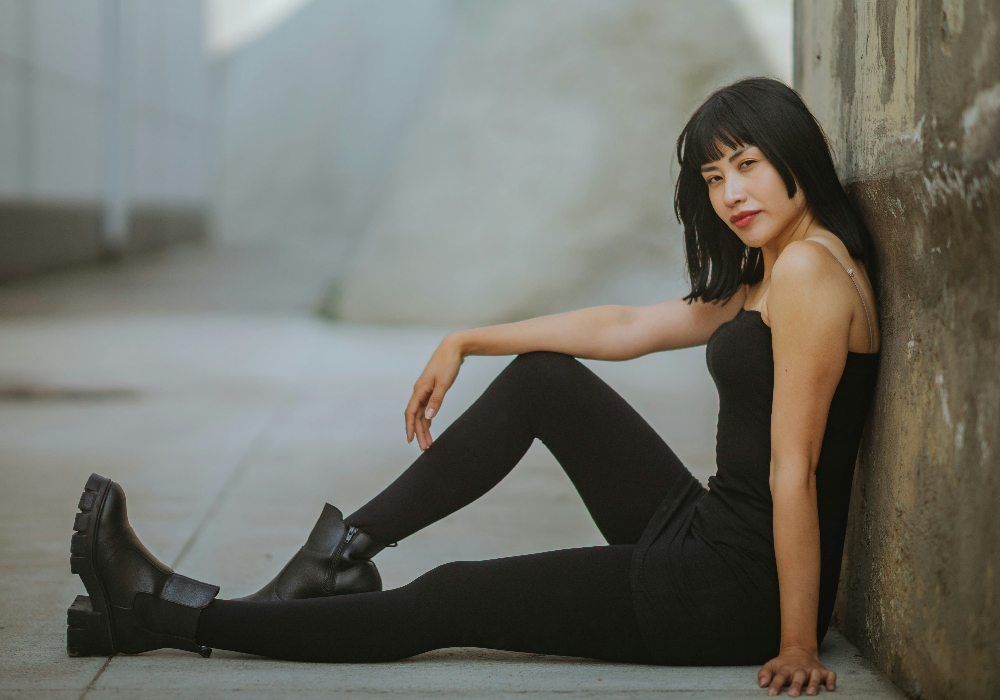 Woman in black minimalist outfit sitting against a wall in a relaxed, casual pose.